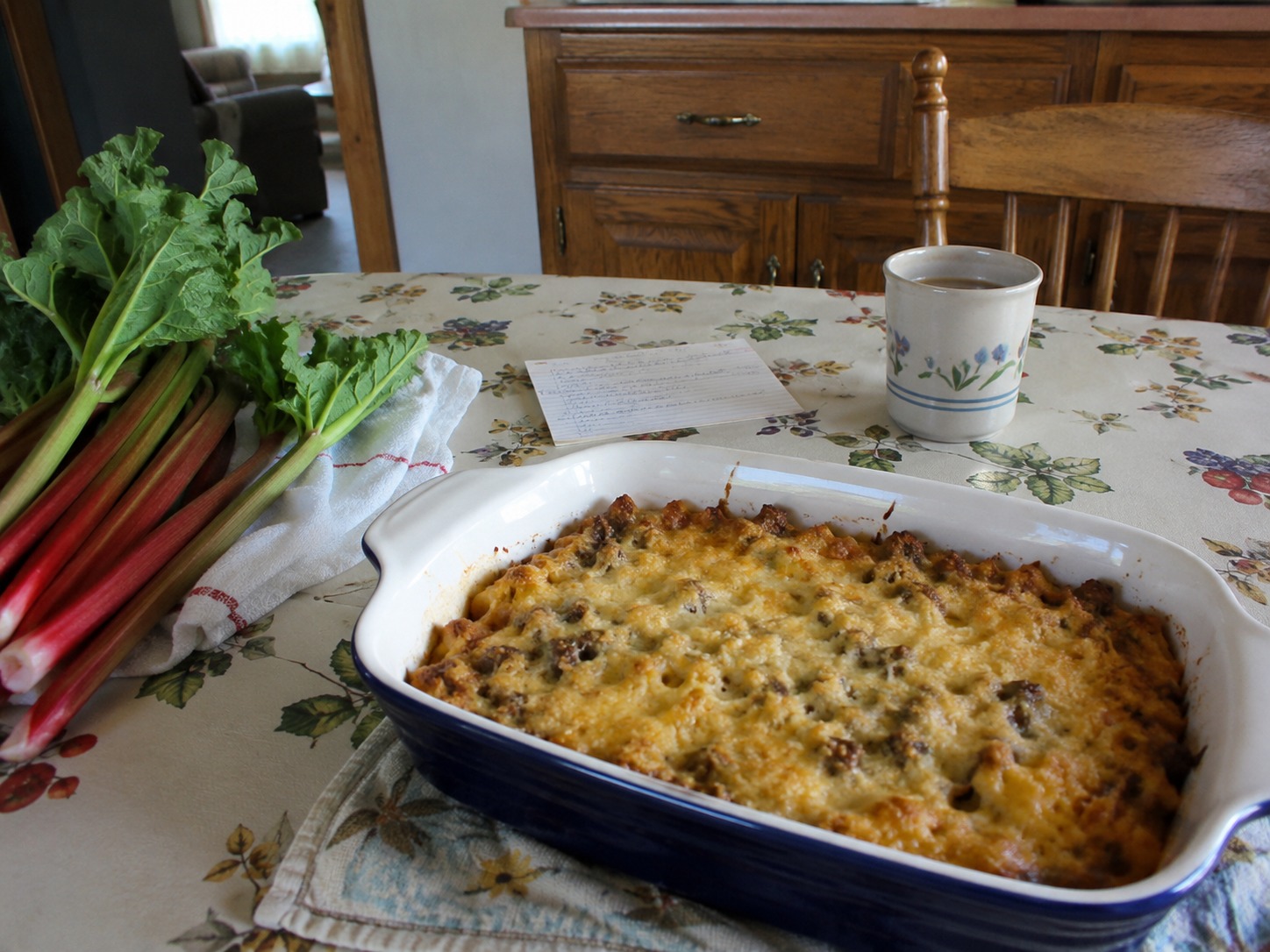 Fresh rhubarb and a casserole dish sit on a kitchen table beside a handwritten recipe card.